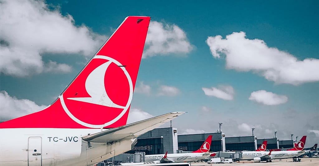 Turkish Airlines plane tail with a busy airport background under a bright blue sky.